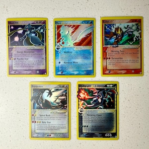 Puede incluir: Una colección de cinco cartas coleccionables rectangulares con ilustraciones de Mewtwo, Mew, Entei, Rayquaza y Charizard. Cada carta muestra arte del personaje, valores de HP, nombres de ataques y texto en inglés, con un borde amarillo.