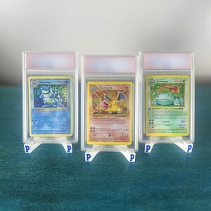 May include: Three collectible trading cards featuring Blastoise, Charizard, and Venusaur, each encased in a clear protective plastic slab. The cards are displayed upright on individual white plastic stands marked with a blue letter P.