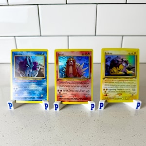 May include: Three rectangular trading cards featuring Suicune, Entei, and Raikou, each displayed in a white plastic stand marked with the letter P. The cards show colorful illustrations, text, and holographic details, standing upright on a flat surface.