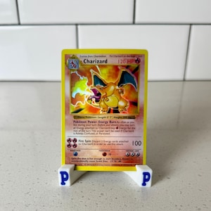 May include: A Charizard trading card featuring a yellow border, a fire-breathing dragon illustration, and 120 HP text. The card is displayed on a small white plastic stand with a blue letter P on each side, set against a white tiled background.