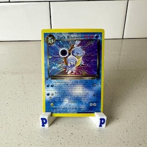 May include: A Dark Blastoise trading card with a holographic illustration of the blue turtle-like creature, featuring 70 HP, Hydrocannon and Rocket Tackle attacks, displayed on a white plastic stand with blue P letters on the front feet.