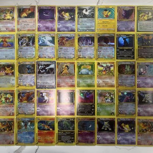 Può includere: Una collezione di quarantadue carte collezionabili rettangolari disposte in una griglia, che presentano varie creature illustrate colorate con testo stampato, statistiche e simboli su ogni carta, inclusi nomi come Charizard, Pikachu e Gengar.
