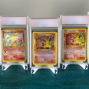 Può includere: Tre carte collezionabili raffiguranti il personaggio Charizard, ciascuna inserita in una custodia protettiva in plastica trasparente con supporto bianco. Le carte presentano illustrazioni colorate, testo e statistiche di gioco.