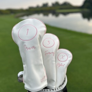 May include: A set of three white golf club head covers with pink piping and embroidery. The covers are labelled 1 Driver, 3 Fairway, and X Hybrid, with each number enclosed in a pink circle. The covers feature the text Caddie Queen in small pink script.