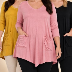 May include: A collection of three v-neck tunics with three-quarter length sleeves and two front patch pockets. The tunics are shown in mustard yellow, dusty rose, and black, each featuring an asymmetrical hemline and a soft, solid-colored fabric.