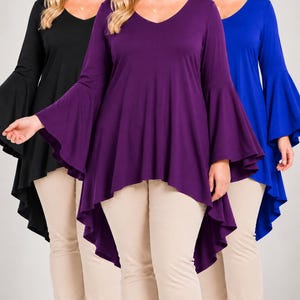 May include: A long-sleeved tunic top with a V-neckline and flared bell sleeves, featuring a high-low hemline. The garment is shown in three solid colors: black, purple, and blue, paired with beige trousers. A small logo reads d2bPLUS.
