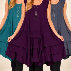 May include: A sleeveless tunic top featuring a round neckline and a multi-layered, handkerchief-style hemline. The garment is shown in teal, deep purple, and dark gray, paired with a long silver-tone necklace. A logo with the text d2bPLUS is visible.