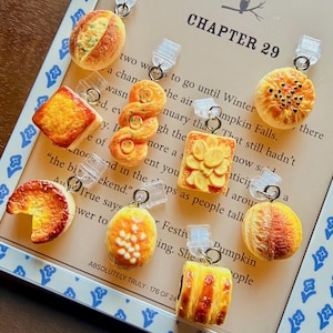 May include: A collection of nine miniature bread and pastry charms, each featuring a clear plastic clip and metal ring. The charms are shaped like various baked goods, including rolls, danishes, and tarts, with realistic golden-brown textures.