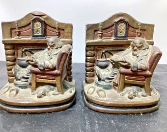 May include: A pair of painted bookends featuring a bearded figure sitting in an armchair reading a book in front of a fireplace. A small dog rests on the floor near the chair, with a cauldron hanging in the hearth and a clock on the mantel.