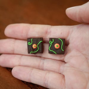 May include: Two square-shaped brown brownie-style charms featuring a dark chocolate-colored top, a small orange pumpkin-shaped decoration in the center, and thin, wavy green icing lines drizzled across the surface of each piece.