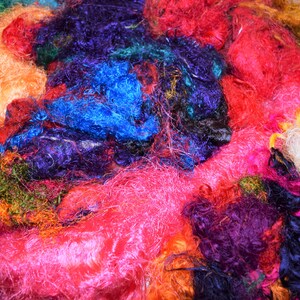 Op de afbeelding: Een stapel kleurrijke, pluche en textuurrijke vezels in tinten roze, rood, blauw, groen, geel en wit. De vezels zijn in een willekeurig patroon gerangschikt, wat een visueel aantrekkelijke en tastbare ervaring cre&euml;ert.