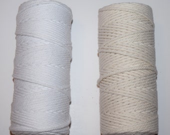 May include: Two spools of macrame cord. One is bright white, the other is a natural off-white color. The cords are tightly wound around cardboard tubes. The texture of the cord is visible, suggesting a soft, yet durable material.