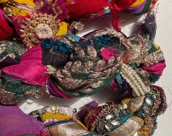 May include: Close-up of colorful braided fabric with intricate embellishments. The fabric features vibrant hues of pink, yellow, red, and green, adorned with gold and silver metallic threads, sequins, and beads. The texture is rich and detailed, suggesting a handcrafted item.