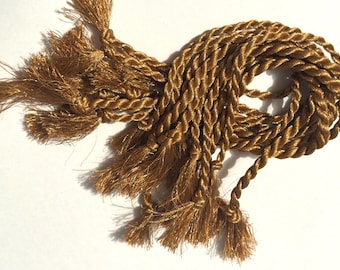 May include: Close-up of a golden-brown decorative rope with multiple tassels. The rope is tightly twisted, and the tassels are frayed at the ends. The rope is likely used for crafting or embellishment.