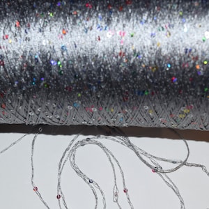 May include: A spool of silver thread with colorful sequins woven throughout. The thread is unwound and draped on a white surface.