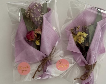 May include: Two small bouquets of dried flowers wrapped in lavender fabric and clear plastic. Each bouquet is tied with twine and has a pink sticker that says "Thank you" in gold lettering. The flowers include roses and other dried blooms.