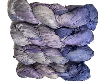 May include: A close-up of a skein of hand-dyed silk ribbon yarn in shades of blue, purple, and gray. The yarn is twisted and has a textured appearance, ideal for fiber art projects.