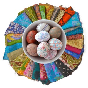 May include: A white bowl filled with decorated Easter eggs sits atop a colorful arrangement of fabric scraps. The eggs feature various patterns and colors, while the fabric scraps display a variety of vibrant hues and designs.