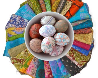 May include: A white bowl filled with decorated Easter eggs sits atop a colorful arrangement of fabric scraps. The eggs feature various patterns and colors, while the fabric scraps display a variety of vibrant hues and designs.