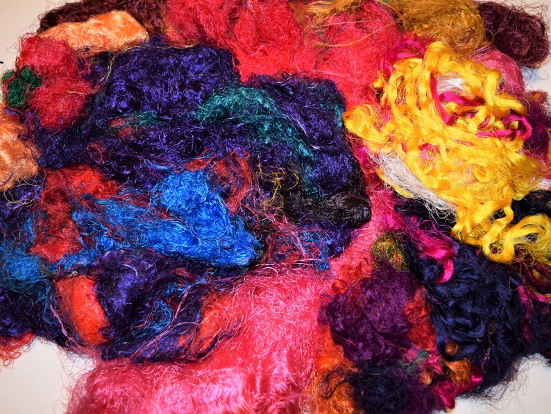 Op de afbeelding: Een stapel kleurrijke zijdevezels in tinten rood, roze, paars, blauw, groen, geel en oranje. De vezels zijn zacht en pluizig en perfect voor knutselen of kunstprojecten.