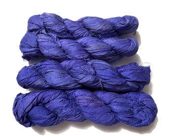 May include: Three skeins of vibrant blue silk ribbon yarn, tightly twisted and arranged against a white background. The yarn appears soft and textured, ideal for various crafting projects. The color is a deep, saturated blue.