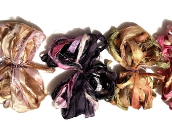 May include: A collection of six silk ribbon bundles in various colors, including shades of pink, purple, gold, and green. Each bundle is tied in the center, creating a flower-like appearance. The ribbons have a slightly textured, crinkled look.