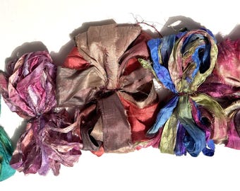 May include: Five colorful silk ribbon bows in shades of green, purple, brown, blue, and pink. The bows are tied in the center and have flowing ribbon ends. The background is white.