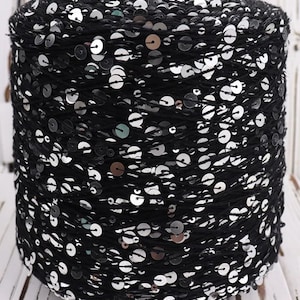 May include: A spool of black yarn densely covered with silver sequins. The yarn is tightly wound, creating a cylindrical shape. The sequins are round and reflective, catching the light and adding a touch of sparkle to the yarn.