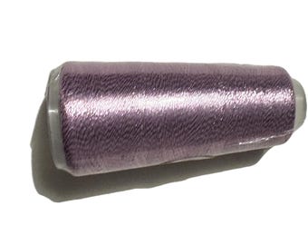 May include: A spool of lavender-colored thread, tightly wound and wrapped in clear plastic. The thread has a subtle sheen, reflecting light. The spool is cylindrical with a white end cap. Ideal for sewing and embroidery projects.