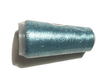 May include: A spool of teal-colored thread, tightly wrapped in clear plastic. The thread has a slight sheen, suggesting a metallic or silk blend. The spool is cylindrical, and the packaging is intact.