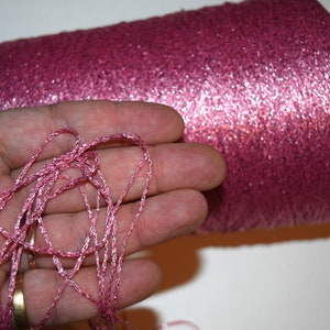 May include: A spool of pink metallic yarn with a strand of yarn in a hand. The yarn has a shimmery finish.