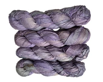 May include: Four bundles of hand-dyed silk ribbon in shades of purple, lavender, and gray, arranged in a braided pattern. The silk has a textured, slightly wrinkled appearance, with subtle variations in color throughout. The background is plain white.