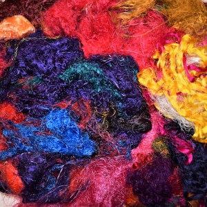 Op de afbeelding: Een stapel kleurrijke zijdevezels in tinten rood, oranje, geel, groen, blauw, paars en bruin. De vezels zijn verward en overlappen elkaar, waardoor een textuur en een abstract patroon ontstaat.