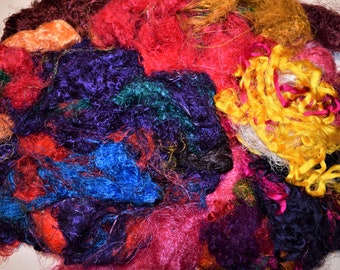 May include: A pile of colorful silk fibers in shades of red, orange, yellow, green, blue, purple, and brown. The fibers are tangled and overlapping, creating a textured and abstract pattern.