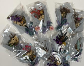 May include: Several small bouquets of dried flowers are wrapped in gray paper and clear plastic. Each bouquet is tied with a purple ribbon and includes a small sticker that says "Thank you". The flowers are various colors, including purple, yellow, and red.