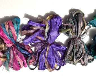 May include: Five bundles of colorful silk ribbon in shades of pink, purple, teal, and green. Each bundle is tied with a thin, dark string. The ribbons have a textured, slightly frayed appearance, suggesting they are handmade.