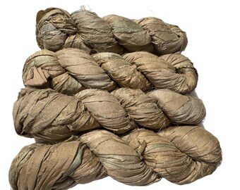 May include: Braided bundles of silk ribbon in shades of tan, beige, and olive green. The silk has a textured, slightly wrinkled appearance. The ribbons are tightly wound and appear to be ready for use in crafting or textile projects.