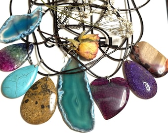 May include: A collection of gemstone pendants in various shapes and colors, including blue, turquoise, purple, and brown. Each pendant is strung on a black cord. A dried rose and small white flowers are also present.