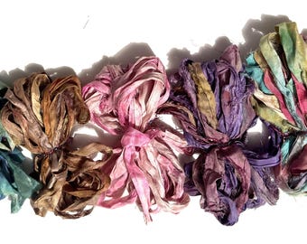May include: A collection of six bundles of colorful silk ribbon. The ribbons are in shades of blue, brown, pink, purple, and multi-colored pastels. Each bundle is tied with a thin string, creating a decorative and textured appearance.