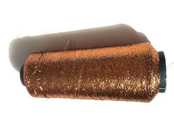 May include: A spool of metallic copper-colored thread. The thread is tightly wound around a dark blue core. The thread has a shiny, glittery appearance, reflecting light. Ideal for embroidery, sewing, or crafting projects.