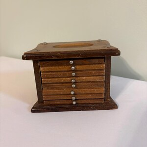 May include: A wooden coaster set housed in a rectangular storage holder. The set includes eight square brown wooden coasters stacked vertically, each featuring a small silver-toned metal knob on the front edge for easy removal from the holder.