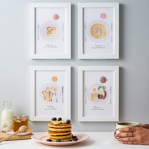 May include: Four white framed prints with illustrations of different breakfast meals. Each print has a title with the name of the person and the text "favorite breakfast".