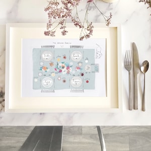 May include: A framed illustration of a family dinner table setting with place cards for each person, including "Freda", "Ross", "Pippa", and "Frank". The illustration is on a light blue background with a white frame.