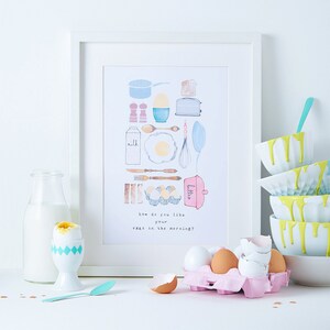 May include: A watercolor illustration of breakfast ingredients, including eggs, milk, butter, and a toaster, with the text "How do you like your eggs in the morning?"  The illustration is framed and displayed on a white wall, with a white table in the foreground. There are several eggs in a carton, a boiled egg in an egg cup, and a glass bottle of milk on the table.