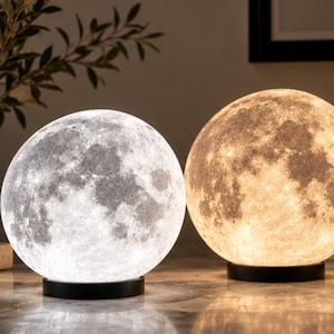 May include: Two spherical moon lamps resting on circular black bases. The left lamp emits a cool white light, while the right lamp glows with a warm yellow light. Both lamps feature a detailed, textured surface replicating the lunar crater landscape.