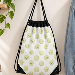 Puede incluir: Una bolsa de cordón blanca con un patrón repetitivo de pequeños personajes de dibujos animados redondos de color verde claro con diversas expresiones faciales. La bolsa tiene cordones negros y esquinas reforzadas negras, colgada de un gancho.