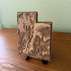 May include: A three-dimensional topographic map of the state of Utah, crafted in layered shades of tan and brown. The relief map features intricate contour lines representing elevation changes and is supported by a small black stand.