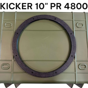 May include: A black circular speaker gasket ring with eight mounting holes, positioned on an olive green rectangular storage case. The text KICKER 10" PR 4800 is printed in black above the object, indicating a 10-inch component for audio equipment.