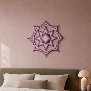 Op de afbeelding: Een wandhanger van garen in een stervormig geometrisch patroon. Het ontwerp bestaat uit concentrische lagen paarse, lavendelkleurige en crèmekleurige draad gewikkeld rond een frame, wat een symmetrische, veelpuntige sterstructuur vormt.
