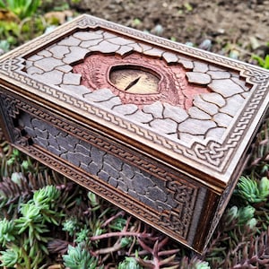 May include: A rectangular wooden box featuring a carved dragon eye at the centre of the lid. The lid and sides are decorated with a cracked stone texture and intricate Celtic knotwork borders. The wood has a dark brown finish with reddish tones.
